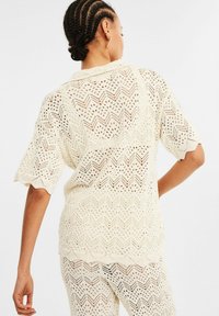 Krämfärgad spetströja i knitting med ett zigzag-mönster, vågig nederkant och korta ärmar, med en öppen väv och texturerad yta.