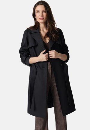 Kobieta poprawia czarny trencz na brązowych spodniach w kratę, patrząc lekko na bok, z luźnymi, falującymi włosami na jasnym tle.