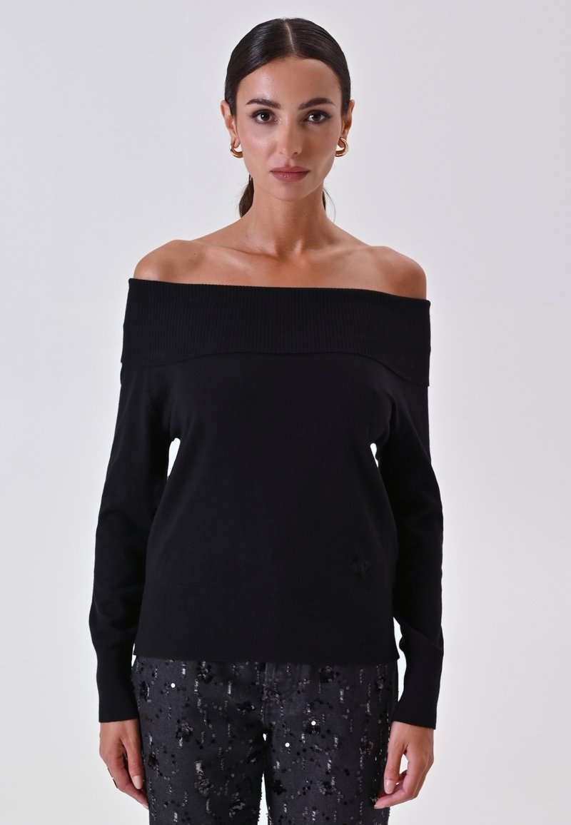 Maglione nero off-the-shoulder con scollatura a costine, maniche lunghe e texture liscia. Abbinato a pantaloni neri con paillettes.