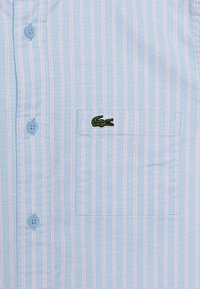 Camicia azzurro chiaro con righe rosa, dotata di taschino e un logo di coccodrillo ricamato in verde sulla tasca.