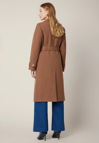 Brązowy wełniany płaszcz typu trench z paskiem w talii, zapinany na guziki rękawami i dopasowanym krojem, zestawiony z szerokimi niebieskimi dżinsami i czarnymi szpilkami.