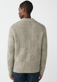 Mężczyzna w beżowym swetrze z dzianiny o teksturowanym kwadratowym wzorze, pokazany od tyłu, w zestawieniu z ciemnoniebieskimi dżinsami na jednolitym tle.