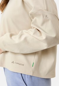 Beige Langarmoberteil mit weicher Textur, das ein dezentes gesticktes Logo und ein kleines grünes Akzentetikett am Saum aufweist.