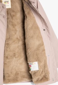 Lyserød jakke med en blød, beige faux fur-foring. Har trykknapper og et mærke med farverigt design, der angiver mærke og størrelse.