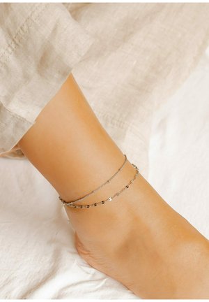 Zilveren enkelbandjes die samen zijn gelaagd, met een mix van delicate kettingpatronen en kleine geometrische accenten tegen een blote voet.
