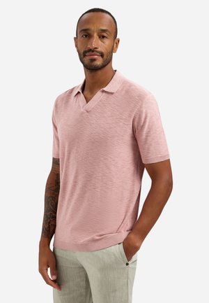 Man met getatoeëerde arm, gekleed in een lichtroze korte mouw gebreide poloshirt en lichtbeige broek, staand met één hand in de zak.