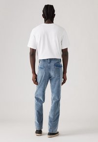 Homme aux cheveux tressés portant un t-shirt blanc, un jean bleu clair et des baskets noires, debout de dos sur un fond uni.