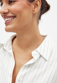 Camicia bianca con strisce verticali azzurre, caratterizzata da una texture morbida, colletto e una patta con bottoni. Primo piano sul colletto.
