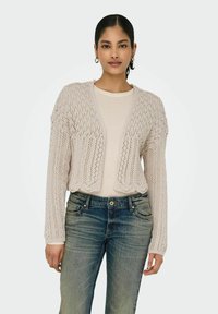 Cardigan lavorato a maglia di un delicato rosa chiaro, con orlo scallop e apertura frontale, caratterizzato da un design in pizzo e maniche lunghe. Indossato sopra una maglia color crema e jeans in denim.