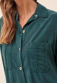 Camicia a maniche lunghe color verde petrolio in tessuto morbido, con due tasche applicate, bottoni color oro rosa e scollo con colletto, che mostra una texture liscia.