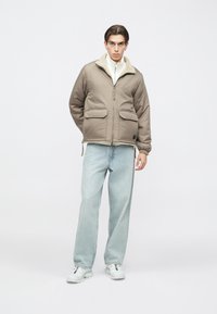 Brun isoleret jakke med en blød hvid krave, to forlommer, kombineret med lyseblå jeans og hvide sneakers, sat op mod en ensfarvet baggrund.