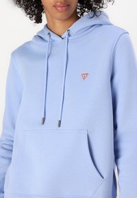 Γαλάζια μπλούζα φούτερ από μαλακό ύφασμα, με μπροστινή τσέπη, κορδόνια με μεταλλικά άκρα και ένα μικρό κόκκινο τρίγωνο λογότυπο.