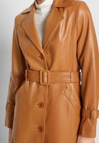 Manteau en cuir camel avec larges revers, boutons croisés et ceinture assortie. Il comprend des poches latérales et des détails texturés.