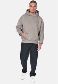 Grå hættetrøje med lomme på forsiden og broderet logo; parret med sorte bukser og hvide sneakers. Modellen står foran en ensfarvet hvid baggrund.