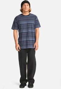 T-shirt com padrão azul-marinho e desenhos geométricos, combinado com calças largas pretas. Modelo a usar sapatos pretos e um gorro de malha.