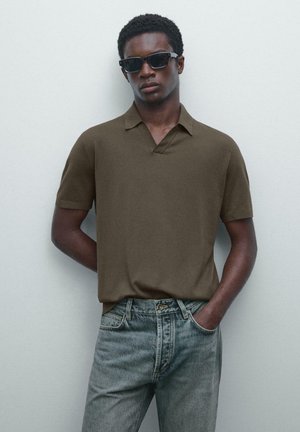 Giovane uomo con occhiali da sole scuri, polo a maniche corte color oliva e jeans azzurri chiari in piedi davanti a una parete bianca semplice.