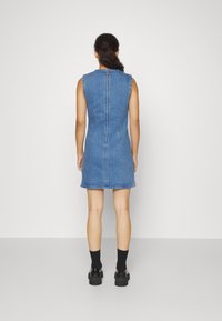 Vestido de mezclilla sin mangas en azul medio con una silueta ajustada, cremallera trasera y dobladillo deshilachado. Combinado con calcetines negros hasta el tobillo y zapatos gruesos.