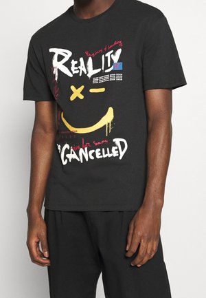 Sort T-shirt med farverig graffiti-stil tekst inklusive "Reality Cancelled", et gult smiley ansigt med X-øjne og en lille blå note med teksten "Have a nice day!".
