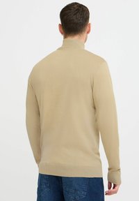 Beige strikkegenser med høy hals, lange ermer og ribbestrikkede mansjetter. Har en glatt tekstur og en kroppsnær silhuett, kombinert med blå jeans.