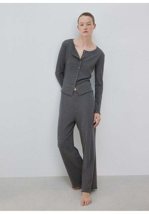 Femme en cardigan gris boutonné et pantalon large assorti, debout pieds nus contre un mur clair uni.