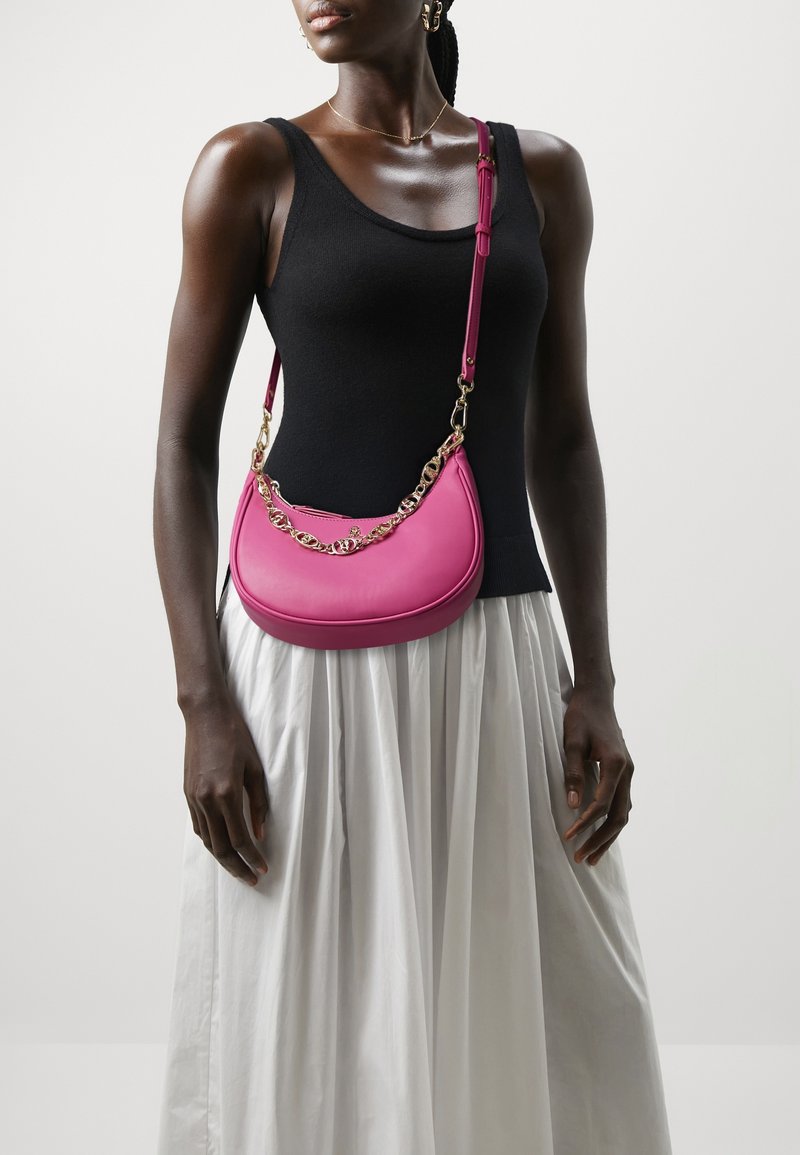 Femme portant un haut sans manches noir et une jupe blanche avec un sac à épaule rose vif orné d'un détail chaîne dorée.