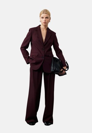 Blazer entallado burdeos con escote en V profundo, combinado con pantalones de pierna ancha. El modelo sostiene un bolso negro y está de pie frente a un fondo neutral.