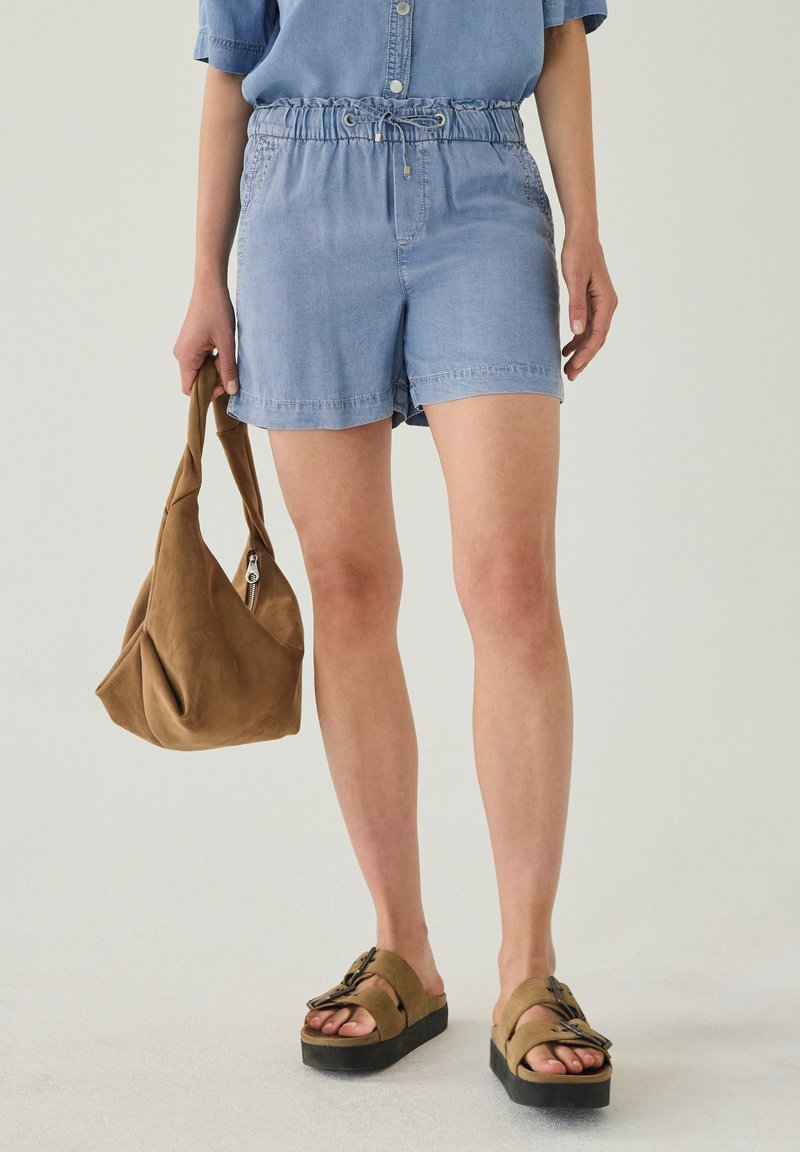 Personne portant un short bleu clair à cordon, tenant un sac à main souple marron, et portant des sandales compensées beige avec boucles.
