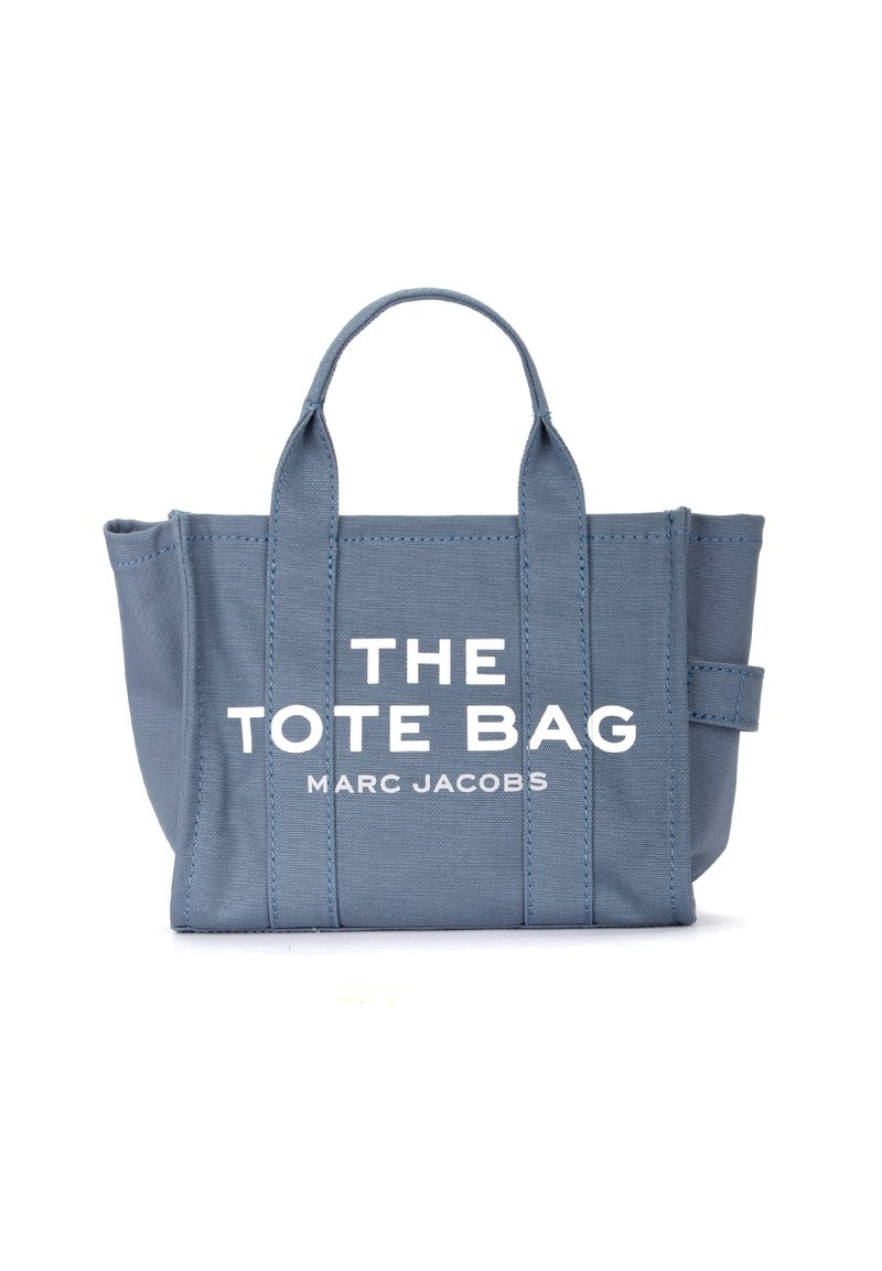 Sac fourre-tout en toile bleu clair avec deux poignées solides, présentant un texte blanc en lettres audacieuses : "LE SAC FOURRE-TOUT MARC JACOBS" à l'avant.