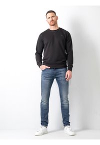 Sudadera de algodón negra con cuello redondo, combinada con jeans de mezclilla azul claro. El modelo se presenta de manera casual con las manos en los bolsillos, usando zapatillas blancas.