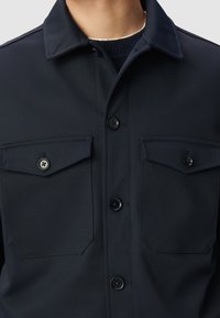 Marineblauwe button-up jas met twee borstzakken, knoopsluiting en een gestructureerde kraag. Gemaakt van een soepele stof met een subtiele textuur.