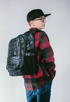 Schwarzer und grauer gemusterter Rucksack mit verstellbaren Trägern, seitlichem Netzfach und glatter Textur. Wird auf dem Rücken getragen, kombiniert mit einem roten Flanellhemd.