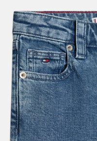Denim jeans detail showing a light blue fabric with a textured finish, top button closure, and a small front pocket featuring a logo patch.