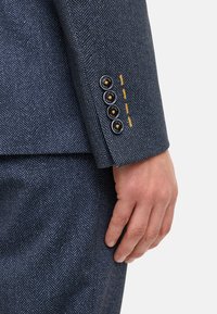 Marine herringbone colbert met contrasterende gele stiksels. Voorzien van vier zwarte knopen op de manchet en een getailleerde pasvorm. Glad textuur.