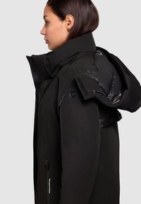 Black insulated jacket with a high collar, zipper, and large hood. Features smooth and textured materials, with a logo on the shoulder strap.