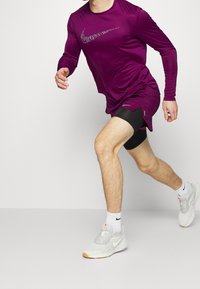 Lila langärmliges Sportshirt mit reflektierendem Nike-Logo, kombiniert mit schwarzen Kompressionsshorts und grauen Laufschuhen.