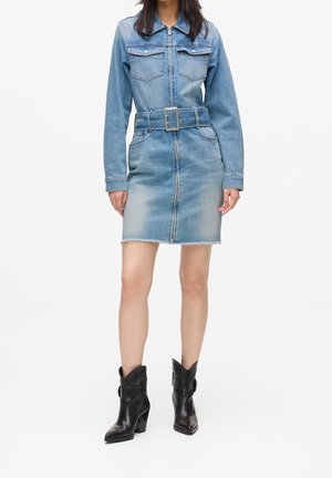 Femme portant une robe en jean bleu clair ceinturée avec manches longues et fermeture éclair à l’avant, associée à des bottines noires en cuir.