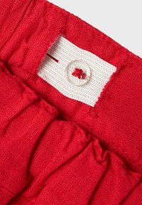 Tissu rouge avec un design froncé, agrémenté d'une étiquette blanche et d'un bouton transparent avec un petit accent rouge. Fini texturé sur l'ensemble.
