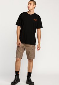 Sort kortærmet t-shirt med orange logo, parret med beige cargo shorts med sidelommer. Modellen har sorte ankelstøvler og sokker på.