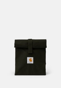Grön canvasväska med ett vikbart lock, säkrat med ett band. Har en vit fyrkantig lapp med en orange logotyp på framsidan.