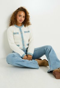 Cardigan blanco suave con detalles en denim azul, cuatro bolsillos, escote redondo y cierre de botones. Combinado con jeans acampanados de color azul claro y botas marrones.