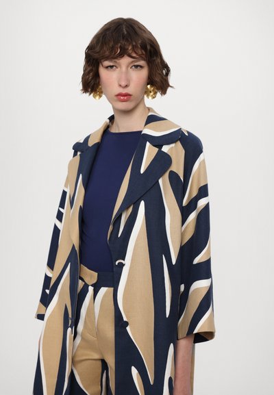 Beige and navy patterned trench coat with a collared neckline, buttons, and wide sleeves, paired with a solid navy top. Gold statement earrings.
