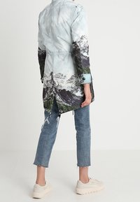 Pitkä, kevyt takki, jossa on vuoristomaisemakuosi sinisissä, harmaissa ja vihreissä sävyissä; yhdistetty lyhennettyihin sinisiin farkkuihin ja vaaleanbeigeihin tennareihin.