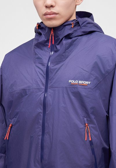Άνδρας που φορά ένα μωβ φούτερ Polo Sport Ralph Lauren με πορτοκαλί φερμουάρ, εμφανίζεται από τους ώμους μέχρι το μέσο του κορμού.