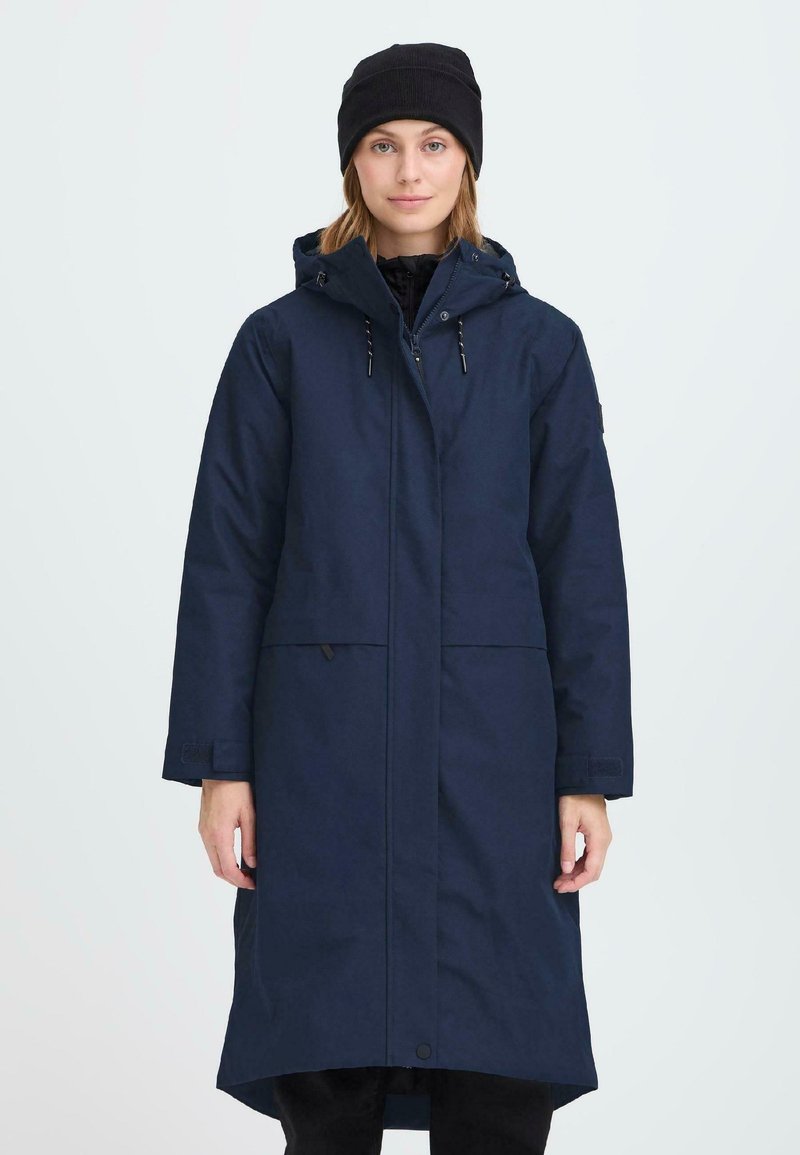 Cappotto lungo navy con cappuccio, dotato di chiusura con zip, due tasche frontali e cordini regolabili. Indossato su una parte inferiore nera e un berretto.