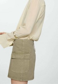 Personne portant un chemisier beige à manches longues avec des poignets évasés et une jupe taille haute kaki dotée d'une grande poche avant.