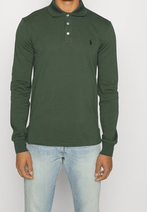 Mann mit einem dunkelgrünen langärmeligen Poloshirt mit drei Knöpfen und hellblauen Jeans, der vor einem einfarbigen Hintergrund steht.