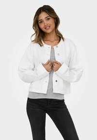 Veste en denim blanche avec un col rond, des boutons pressions argentés et des manches larges, superposée à un haut gris et portée avec un jean noir.