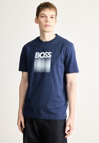 Temno modra bombažna majica z okrolim ovratnikom. Na njej je napis "BOSS" v krepko belih črkah z obliko valov v odtenkih barve spodaj.