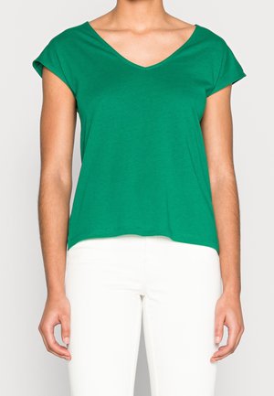 Femme portant un haut vert à col en V et manches courtes, et un pantalon blanc, debout avec les bras détendus le long du corps devant un fond uni.