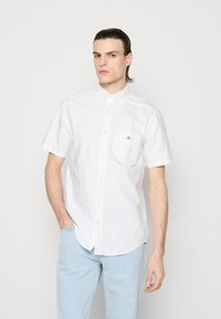 Fiatal férfi, hátrafésült hajjal, fehér rövid ujjú gombos ingben és világoskék farmernadrágban, egy kézzel a zsebében áll.